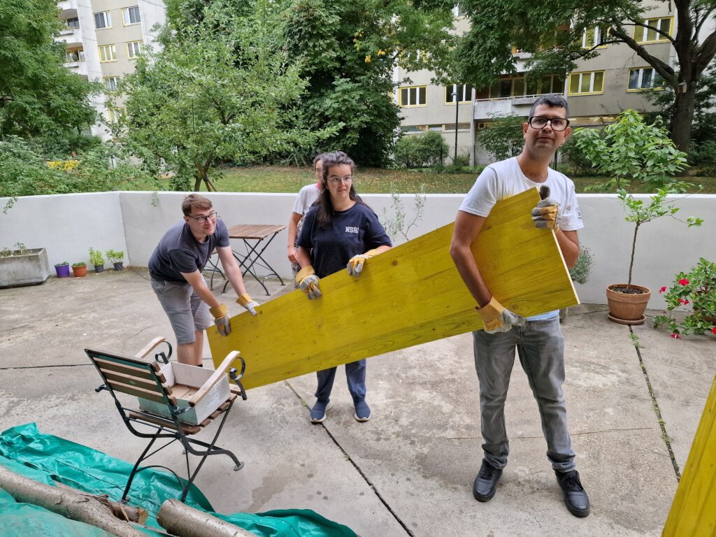 Ein Betreuer trägt eine Holzplatte mit zwei Klient*innen.
