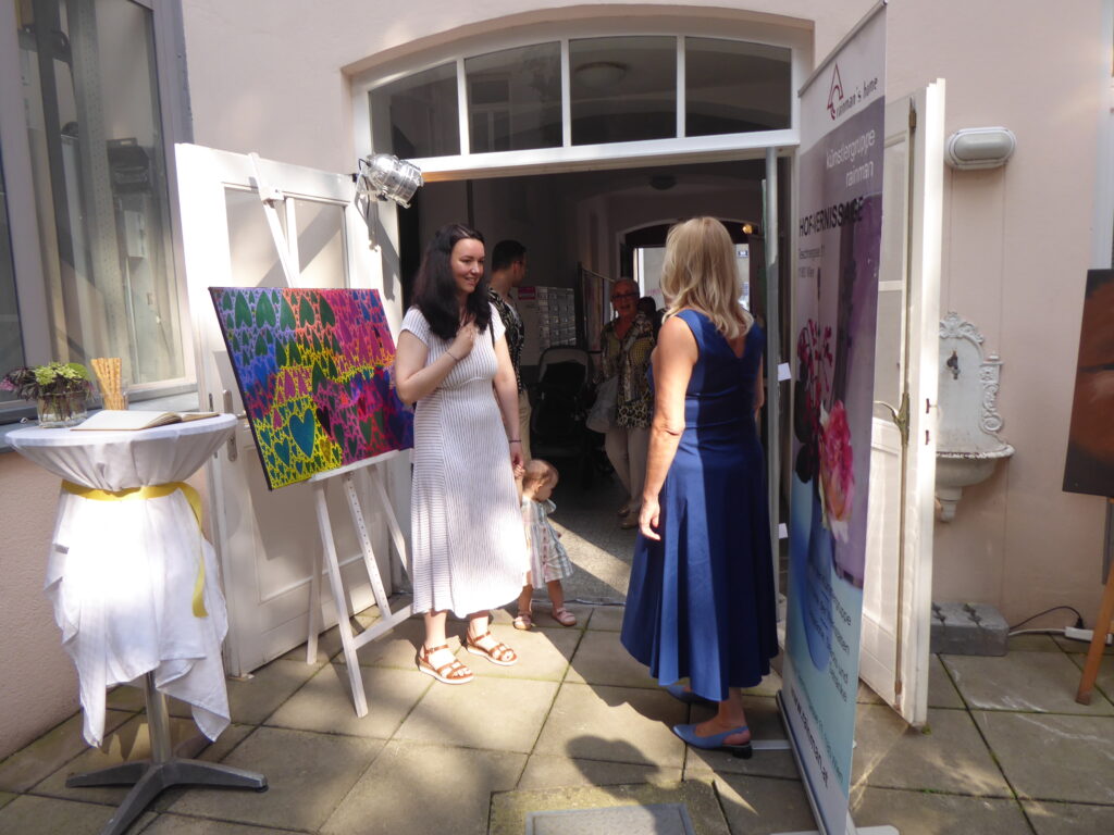 Zwei Frauen welche sich vor dem Eingang der Tagesstruktur unterhalten. Links daneben steht ein gemaltes Bild auf einer Staffelei.