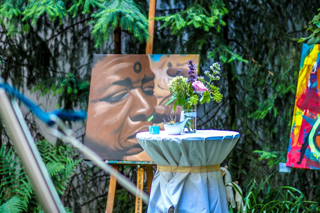 Ein gemaltes Bild einer afrikanischen Person, davor steht ein Tisch mit Blumen.