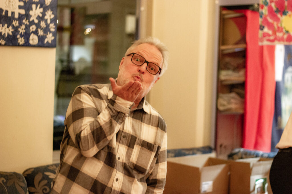 Mittig im Bild ein Mann in karierten Hemd welcher einen Luftkuss in die Richtung der Kamera schickt.