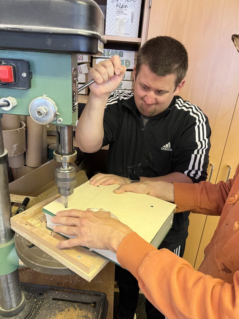 Ein Klient in der Holzwerkstatt bohrt mit einer Standbohrmaschine Löcher in ein Holzbrett.